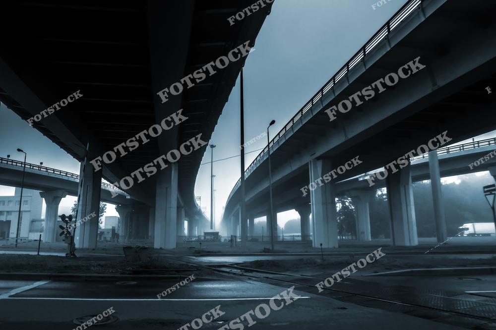 На фотографии изображена одна из мостовых развязок Санкт-Петербурга, погруженная в густой утренний туман. Высокие бетонные опоры и пустая дорога под мостом подчеркивают индустриальную архитектуру города. Туман скрывает дальние объекты, создавая ощущение уединения и загадочности. Этот вид отражает атмосферу спокойствия и безмолвия петербургских улиц в ранние часы.